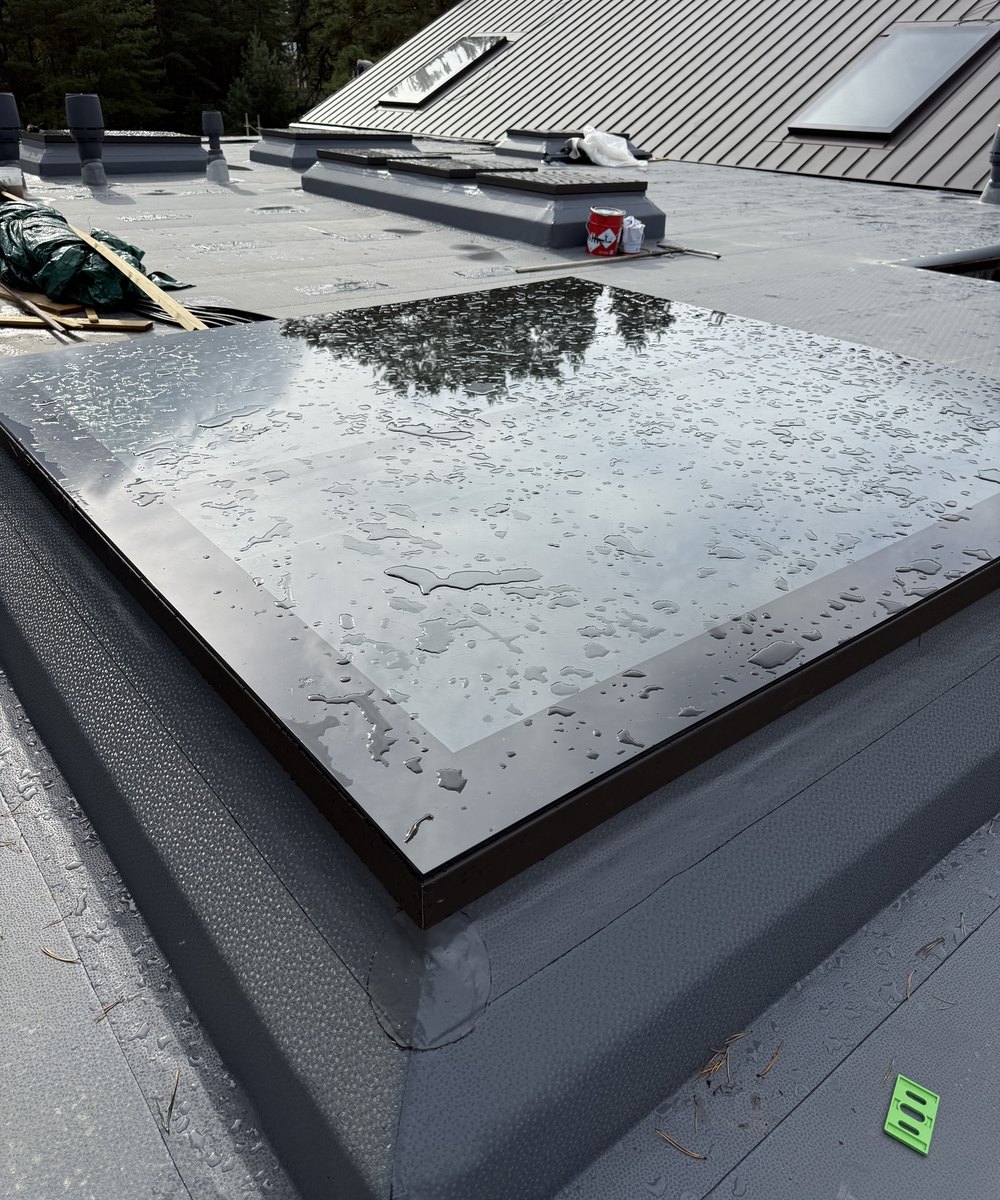 A 2 by 2 metre rooflight on a low-slope membrane roof after rain, pine forest behind